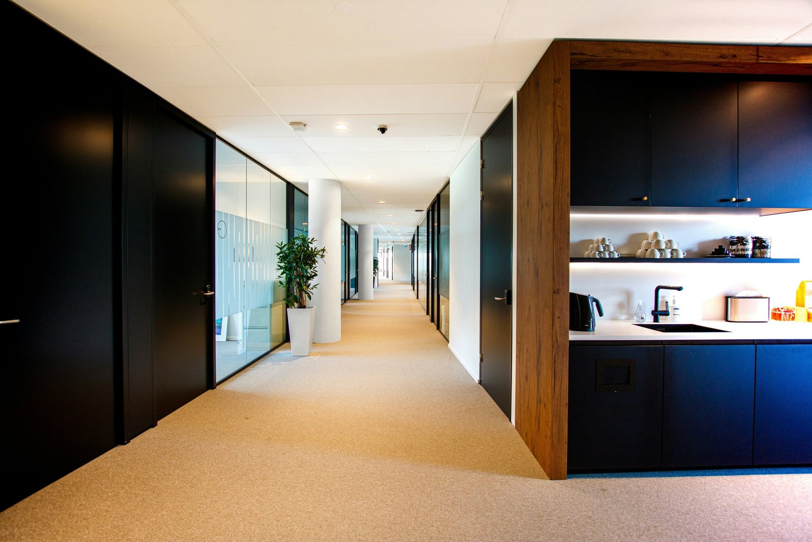 Modern office corridor with glass rooms and a kitchenette.